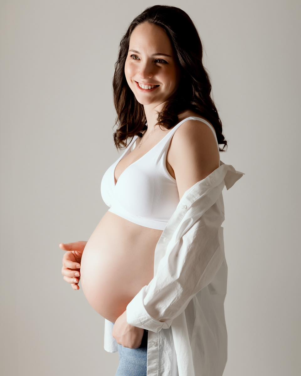 Portrait de femme enceinte souriante en studio, vêtue de blanc, mains sur le ventre, photographe grossesse Annemasse