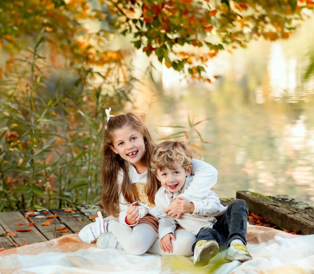 Deux enfants souriants assis au bord de l'eau en automne, feuilles colorées en arrière-plan, photo de famille professionnelle