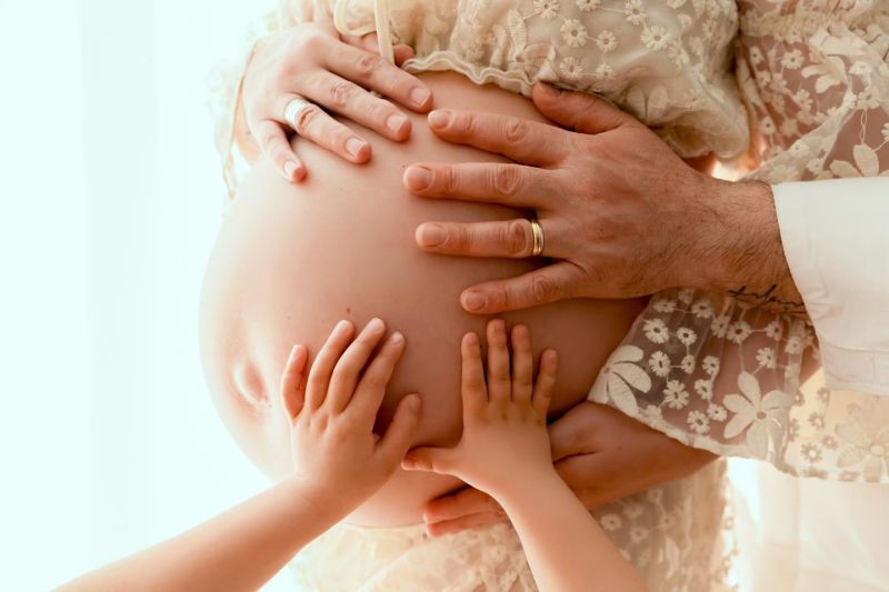 Femme enceinte avec son mari et son enfant posant tendrement les mains sur son ventre arrondi, photographe grossesse à Annemasse, près de Genève