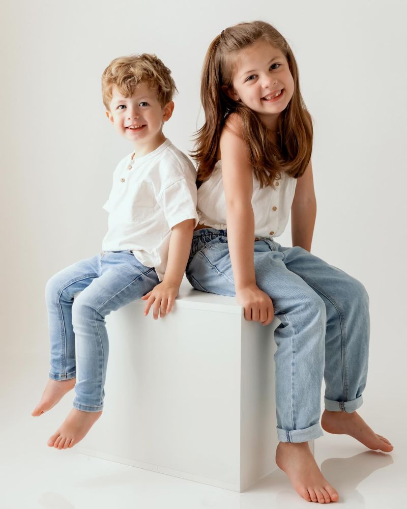 Deux enfants souriants assis sur un cube blanc en studio photo, portant des vêtements blancs et denim, photographe famille à Annemasse