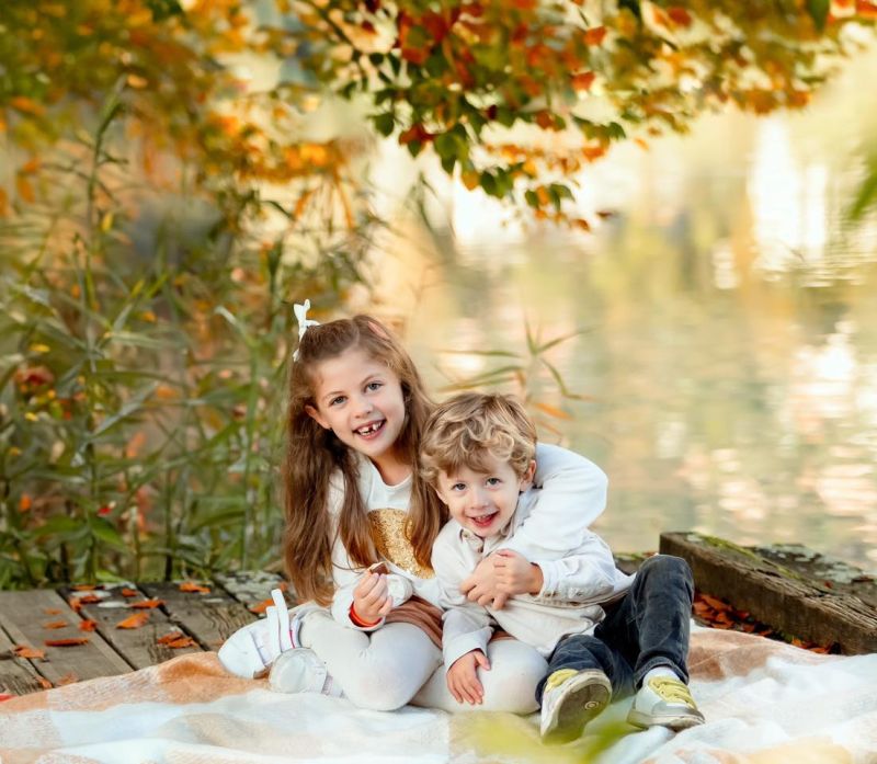 Deux enfants souriants assis au bord de l'eau en automne, feuilles colorées en arrière-plan, photo de famille, photographe famille en Haute-Savoie