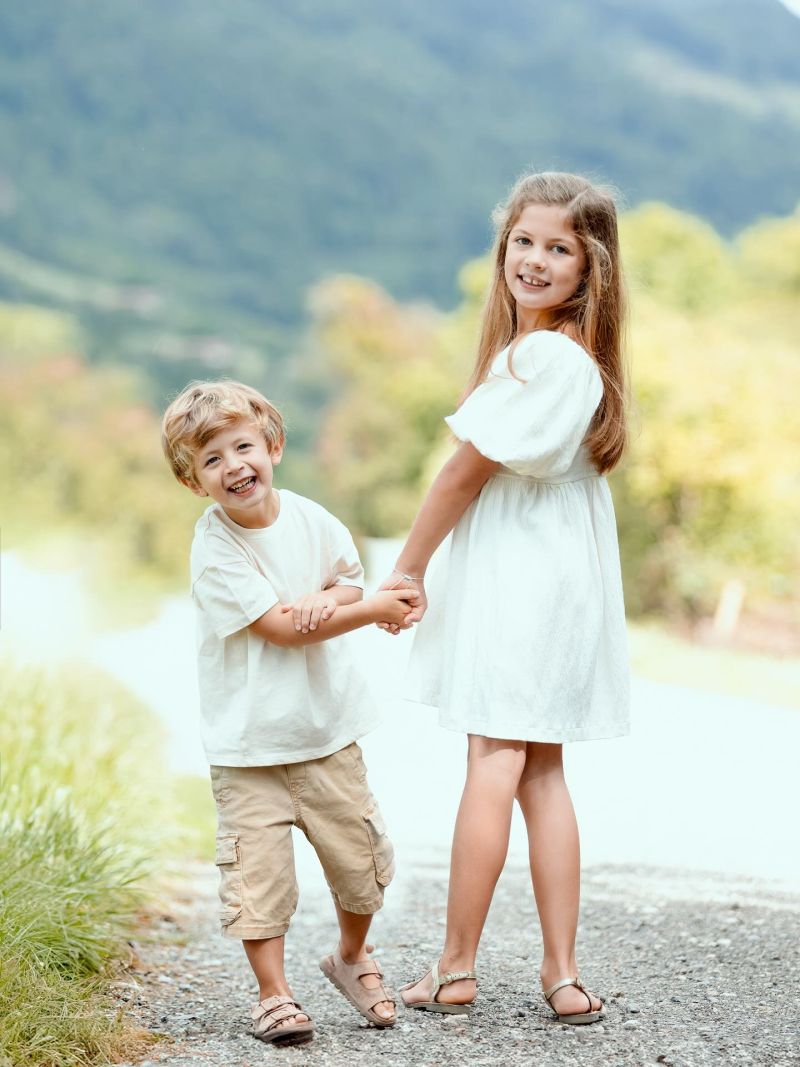 Deux enfants souriants tenant la main sur un chemin de montagne, fond paysage alpin verdoyant, photographe famille à Annemasse