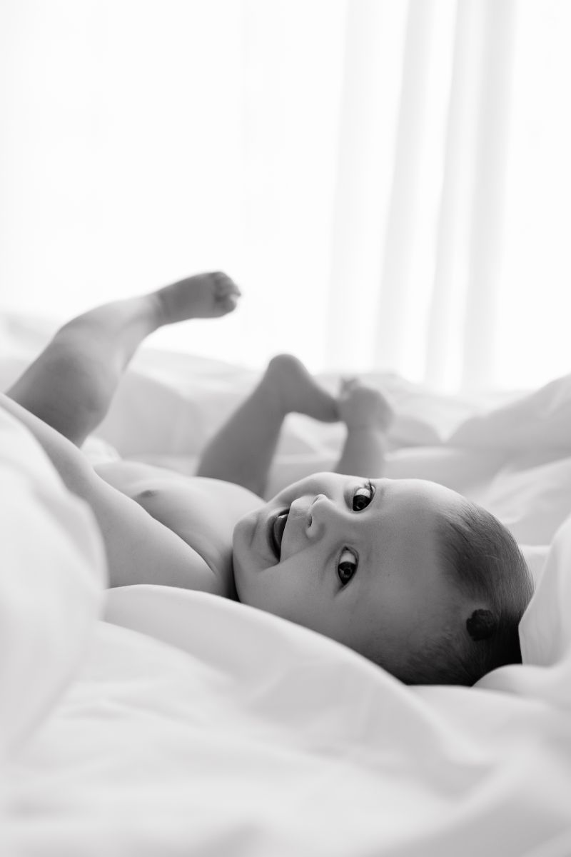 Bébé souriant allongé sur des draps blancs, portrait en noir et blanc réalisé par photographe spécialisée bébé, photographe bébé en Haute-Savoie