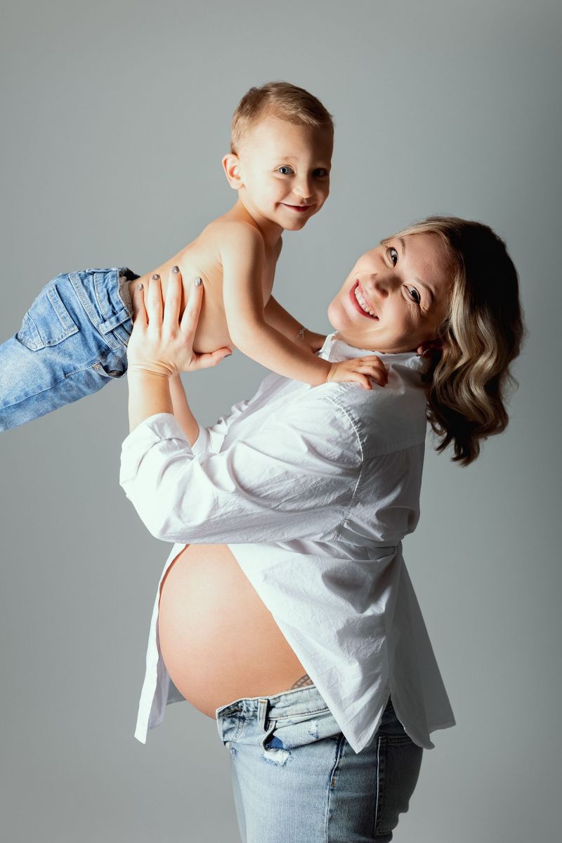 Femme enceinte souriante tenant son enfant en studio, ventre maternel visible, pose tendresse famille, photographe grossesse à Annemasse, près de Genève