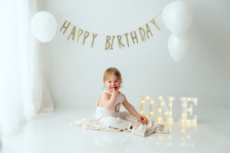 Petite fille souriante de 1 an en robe blanche lors de sa séance photo d'anniversaire au studio, entourée de ballons blancs et d'une décoration dorée « ONE », photographe bébé entre Genève et Annecy