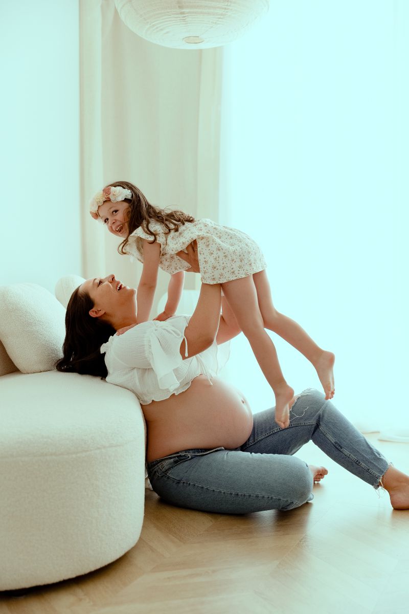 Mère enceinte soulevant sa petite fille sur un canapé blanc en studio, moment de complicité maternelle. Photographe lifestyle près de Genève