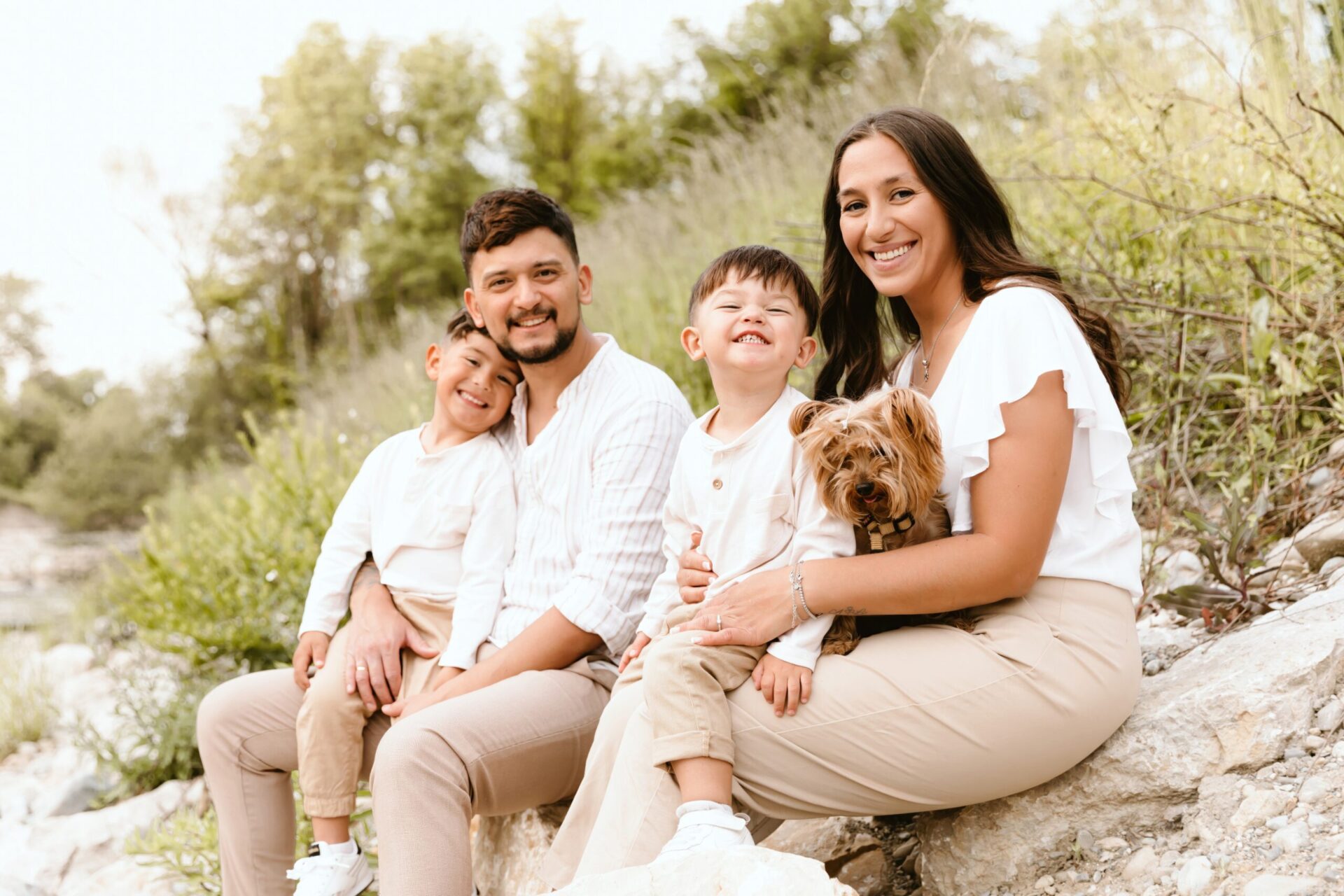 Famille heureuse avec deux enfants et chien en séance photo nature, souriant ensemble en tenue beige et blanc