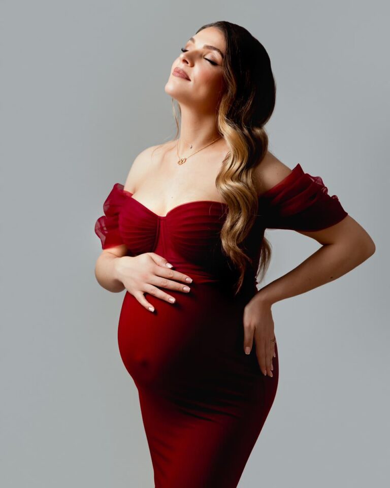 Femme enceinte vêtue d'une robe rouge bordeaux, posant avec délicatesse dans un studio photo professionnel