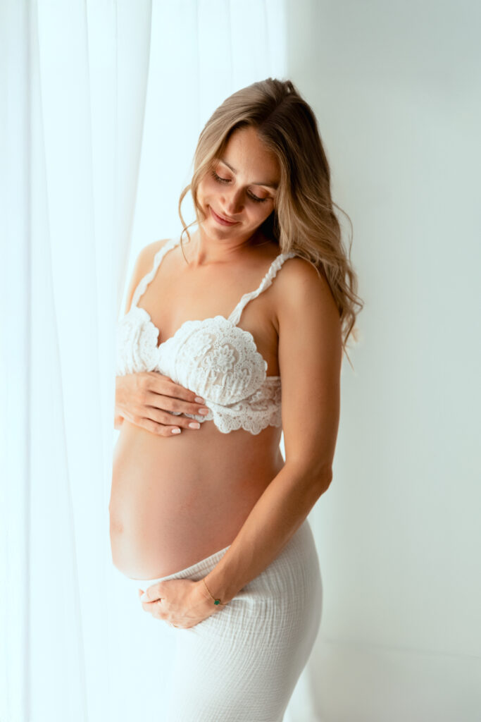 Femme enceinte en séance photo grossesse, vêtue de lingerie blanche, tenant son ventre avec tendresse devant fond blanc lumineux