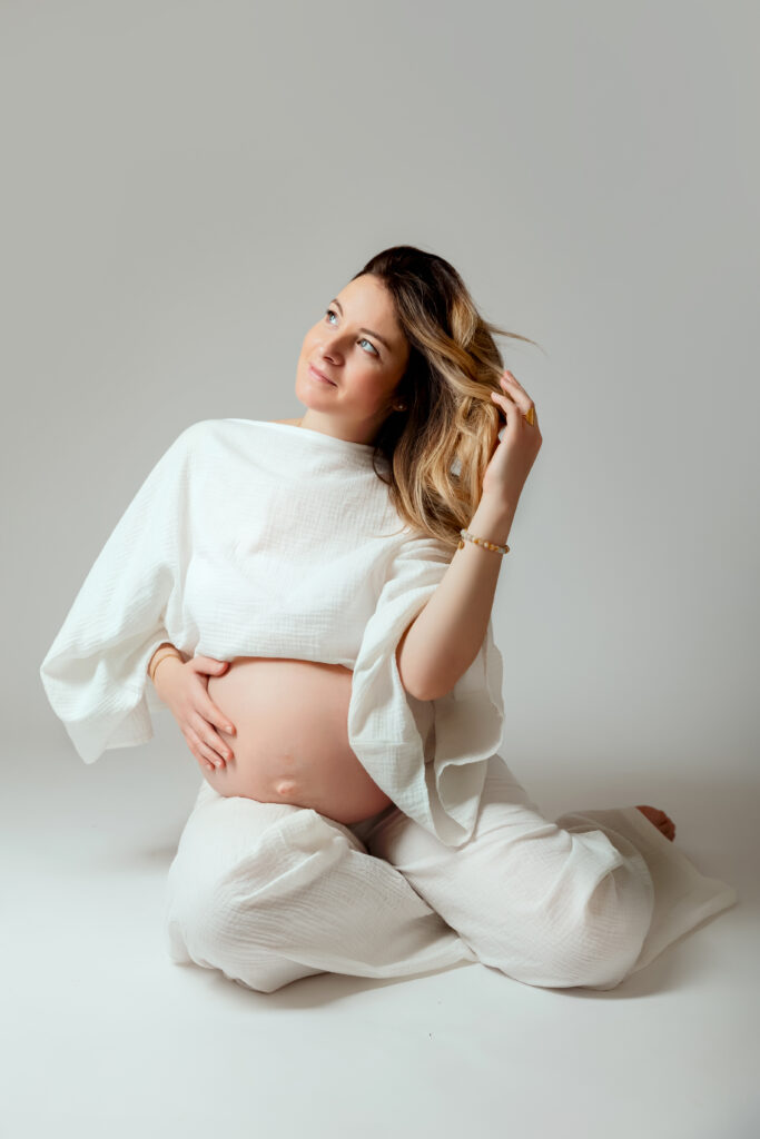 Mum-to-be in white, seated and tenderly holding her bump during a maternity photo session