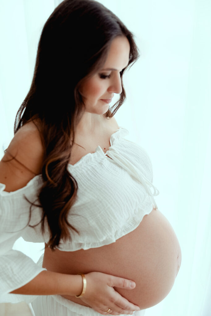 Femme enceinte en robe blanche caressant son ventre, séance photo maternité professionnelle