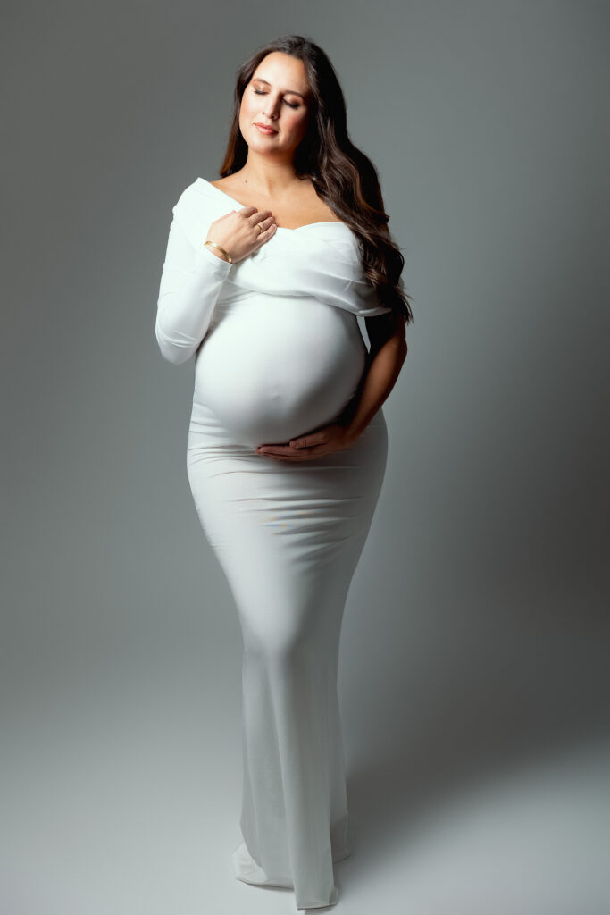 Mum-to-be in a fitted white dress posing in profile, hand on her bump, maternity photographer in Annemasse
