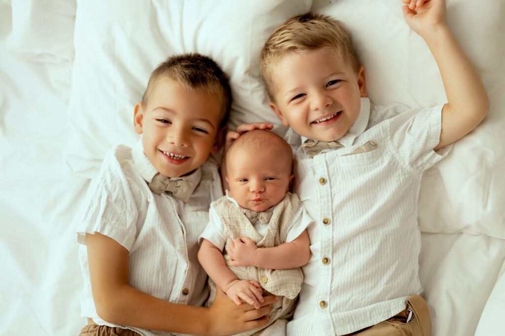 Trois enfants souriants allongés sur un lit blanc, vêtus de vêtements beige clair, frère et sœurs heureux pour séance photo famille