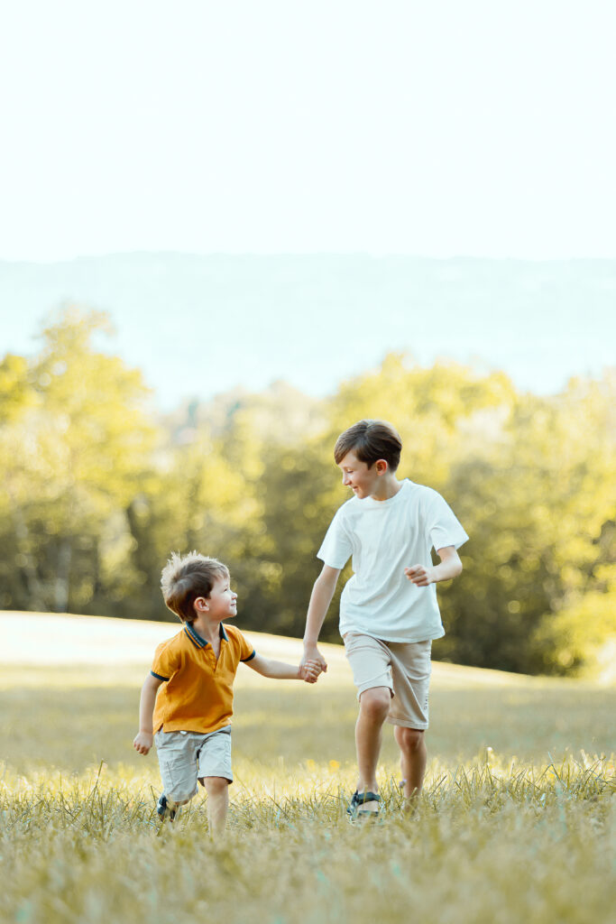 Two children running hand in hand through a sunny field with a forest in the background